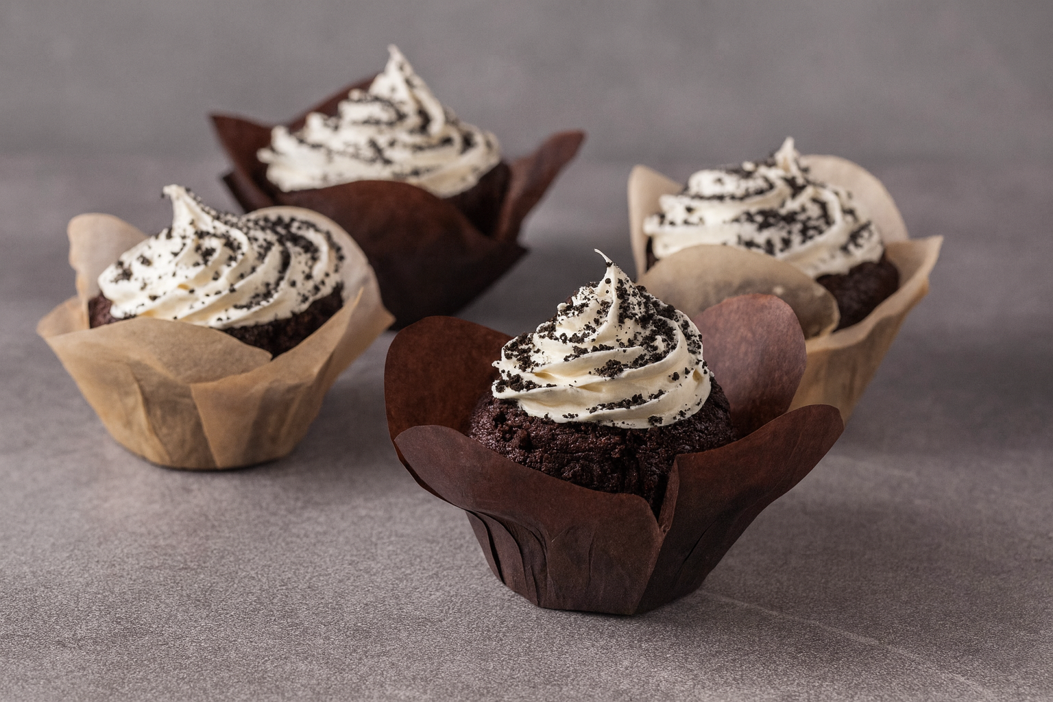 Four chocolate cupcakes with white frosting and cookie crumbles, in brown and beige cupcake liners, on a gray surface.