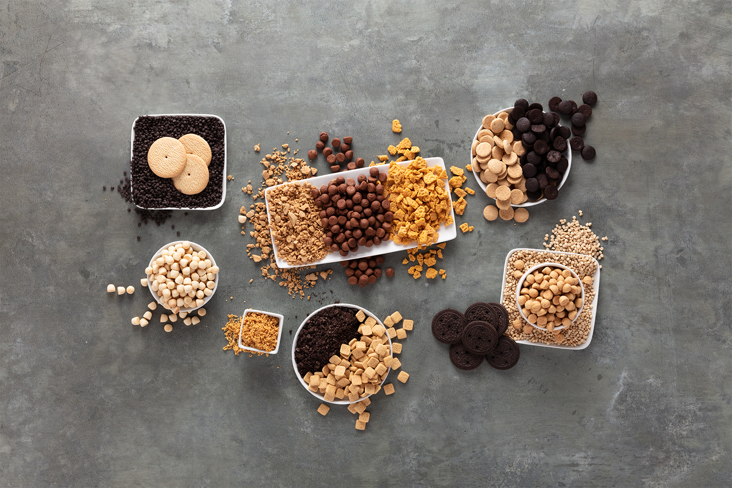 Assorted cookies and baking ingredients on a gray surface, including chocolate chips, cookie dough, crushed cookies, and sprinkles.
