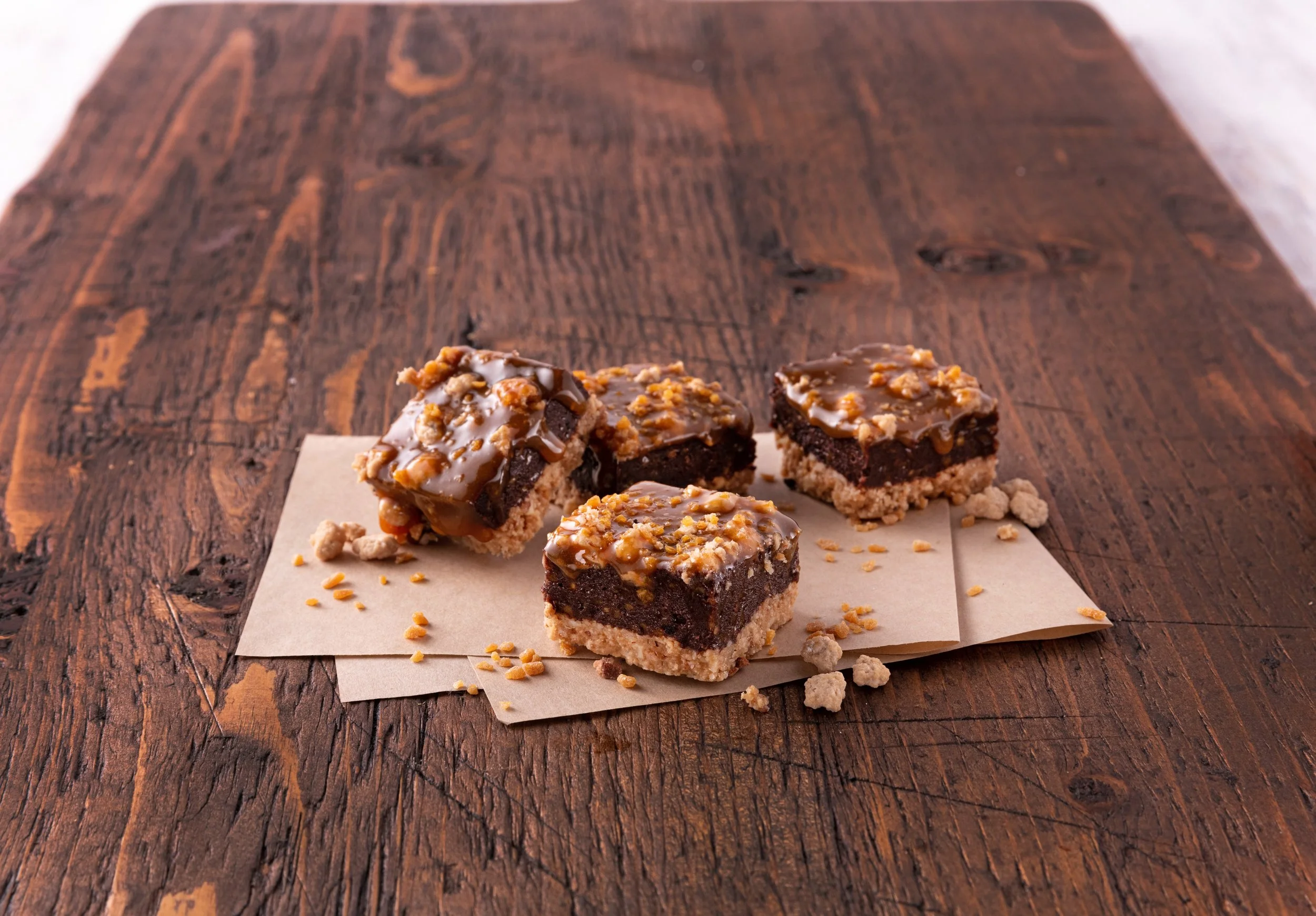 Four brownie squares with a layer of caramel and chopped nuts on top, placed on parchment paper on a wooden surface.