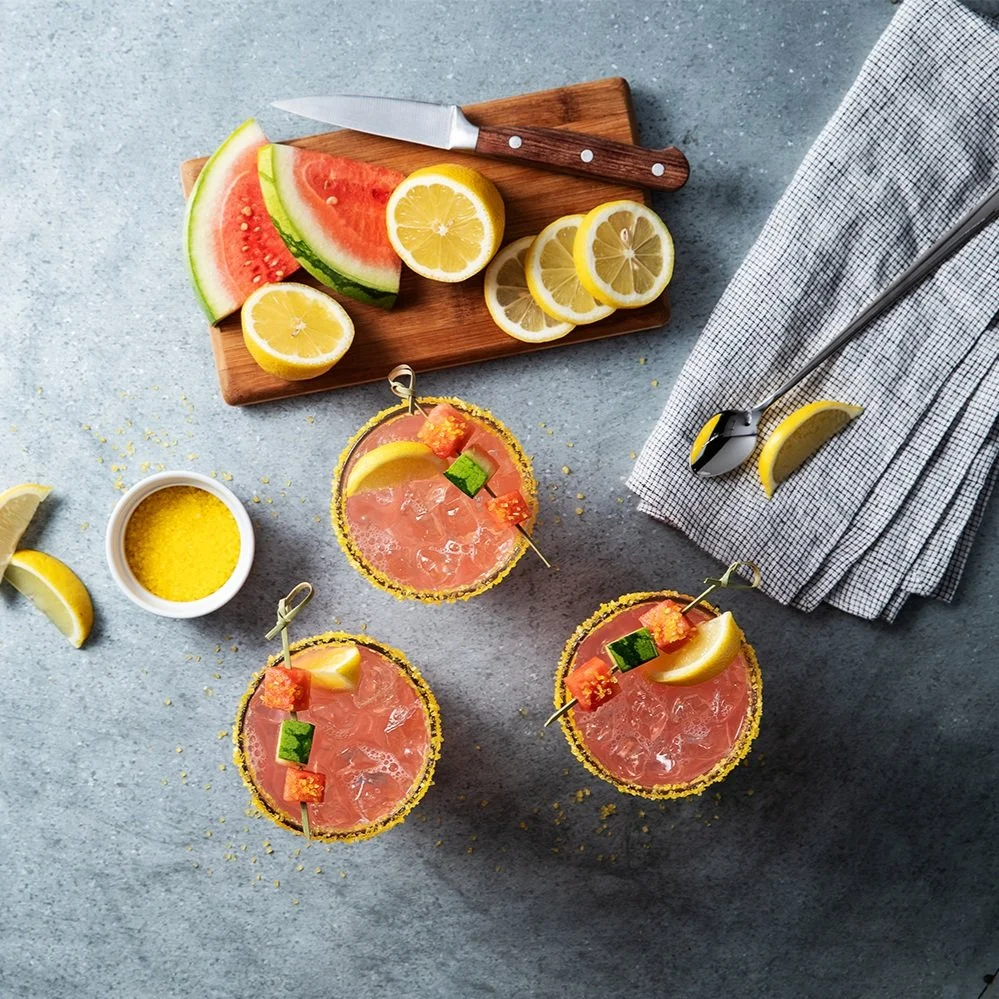 Three pink cocktails garnished with lemon slices and small fruit pieces, with a salted rim, set on a blue surface. A cutting board with watermelon and lemon slices, a small bowl of yellow seasoning, and a napkin with a spoon and lemon wedge are also visible.