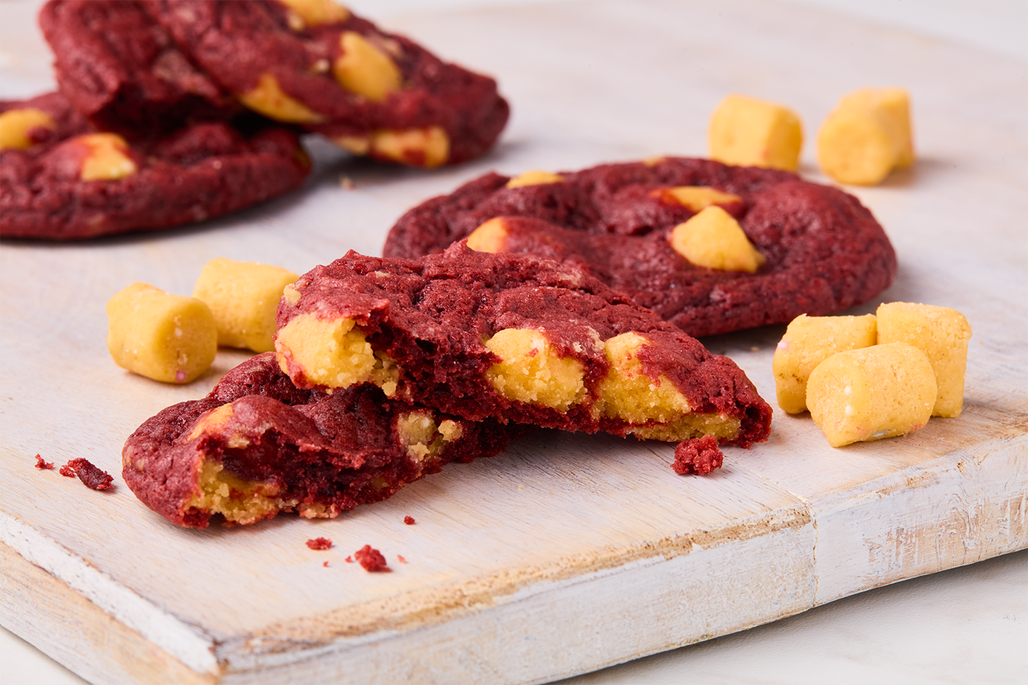 Red velvet cookies with white chocolate chunks on a wooden surface, surrounded by small yellow candy pieces.