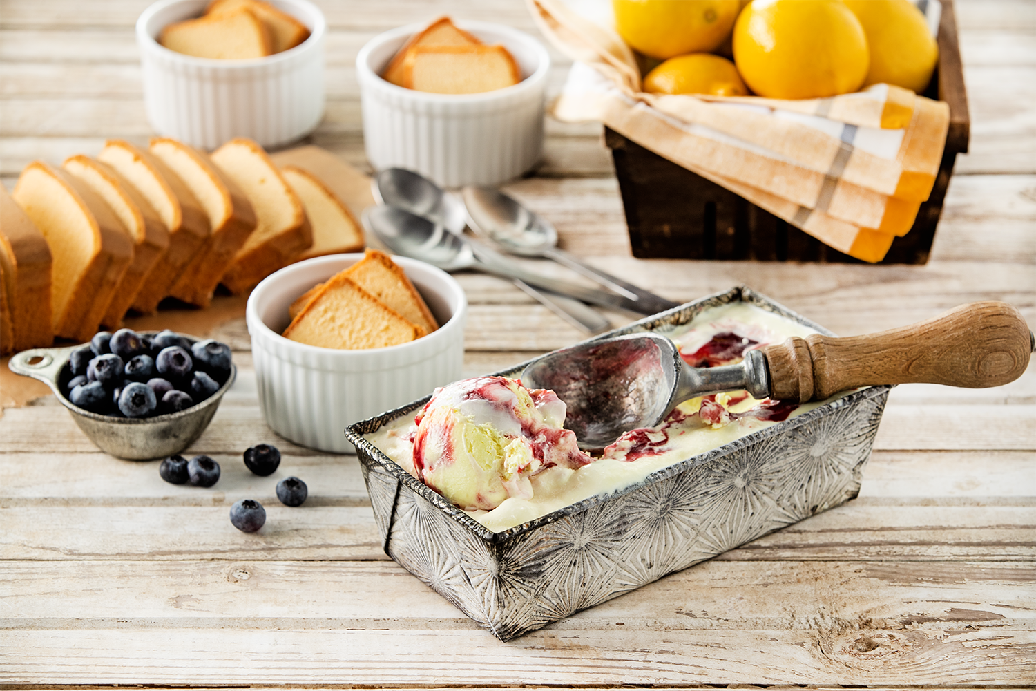 A rustic wooden table with vanilla ice cream topped with raspberry sauce in a decorative metal tray, sliced pound cake, fresh blueberries, and a basket of yellow lemons, with small ceramic ramekins of pound cake slices and a set of silver spoons in the background.