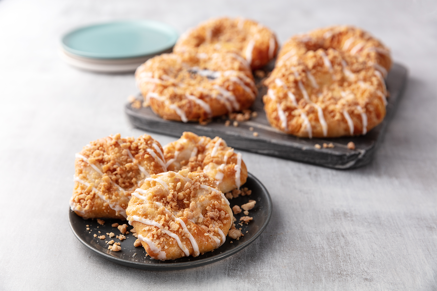Close-up of three glazed donuts with crumb topping on a black plate in the foreground, and five more donuts with similar toppings on a black cutting board in the background, with a stack of small blue plates in the background.