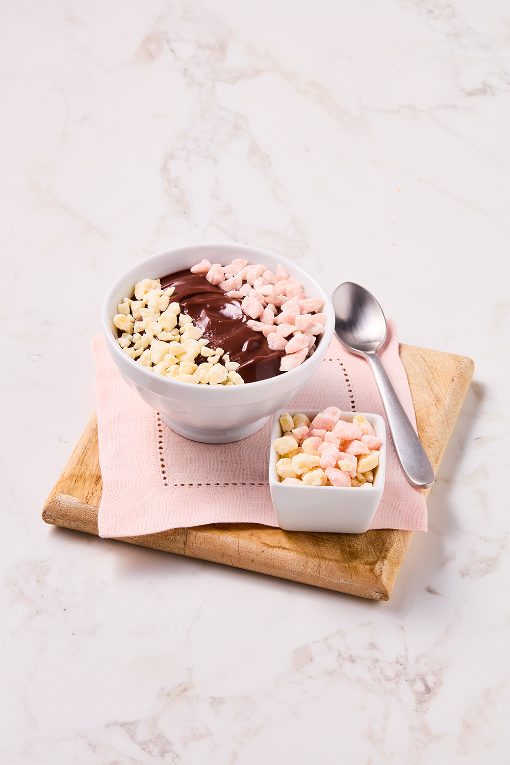 A bowl of chocolate pudding topped with pink and white marshmallows, served on a pink napkin on a wooden board with a small bowl of marshmallows and a metal spoon.