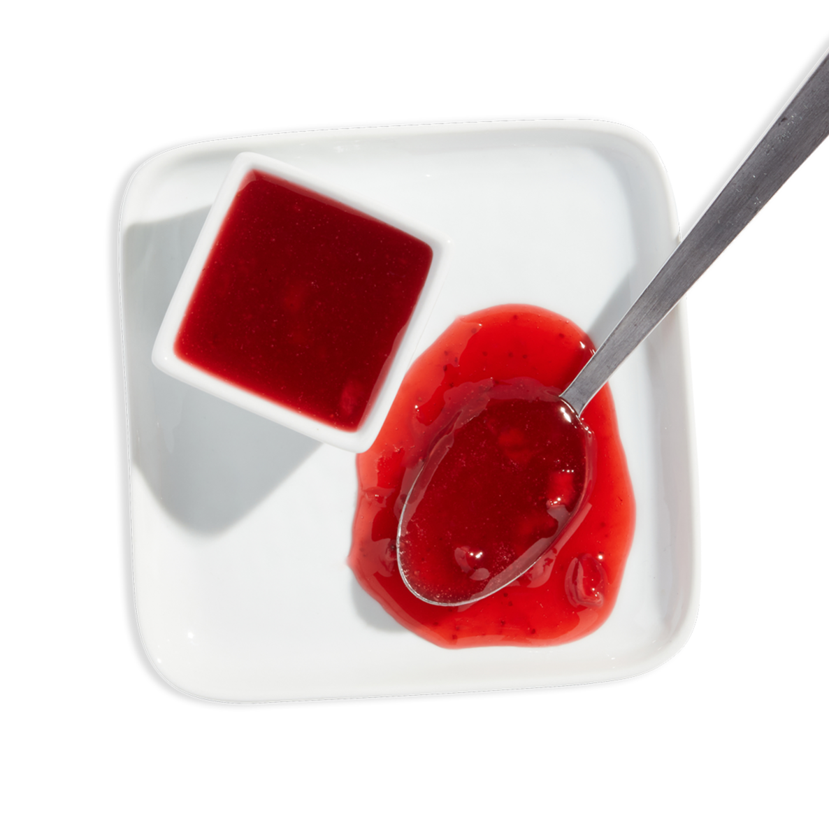 A white square dish containing red raspberry jam and a small bowl of raspberry jelly, with a spoon resting in the jam.