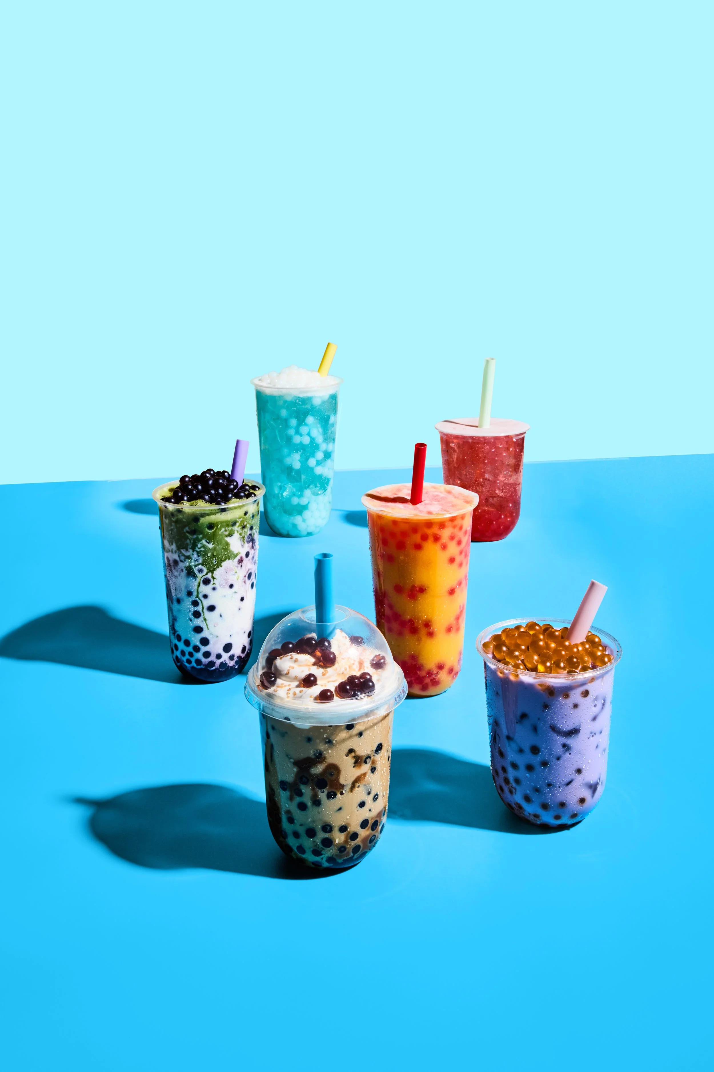 Seven colorful bubble tea drinks with tapioca pearls, arranged on a two-tone blue background.