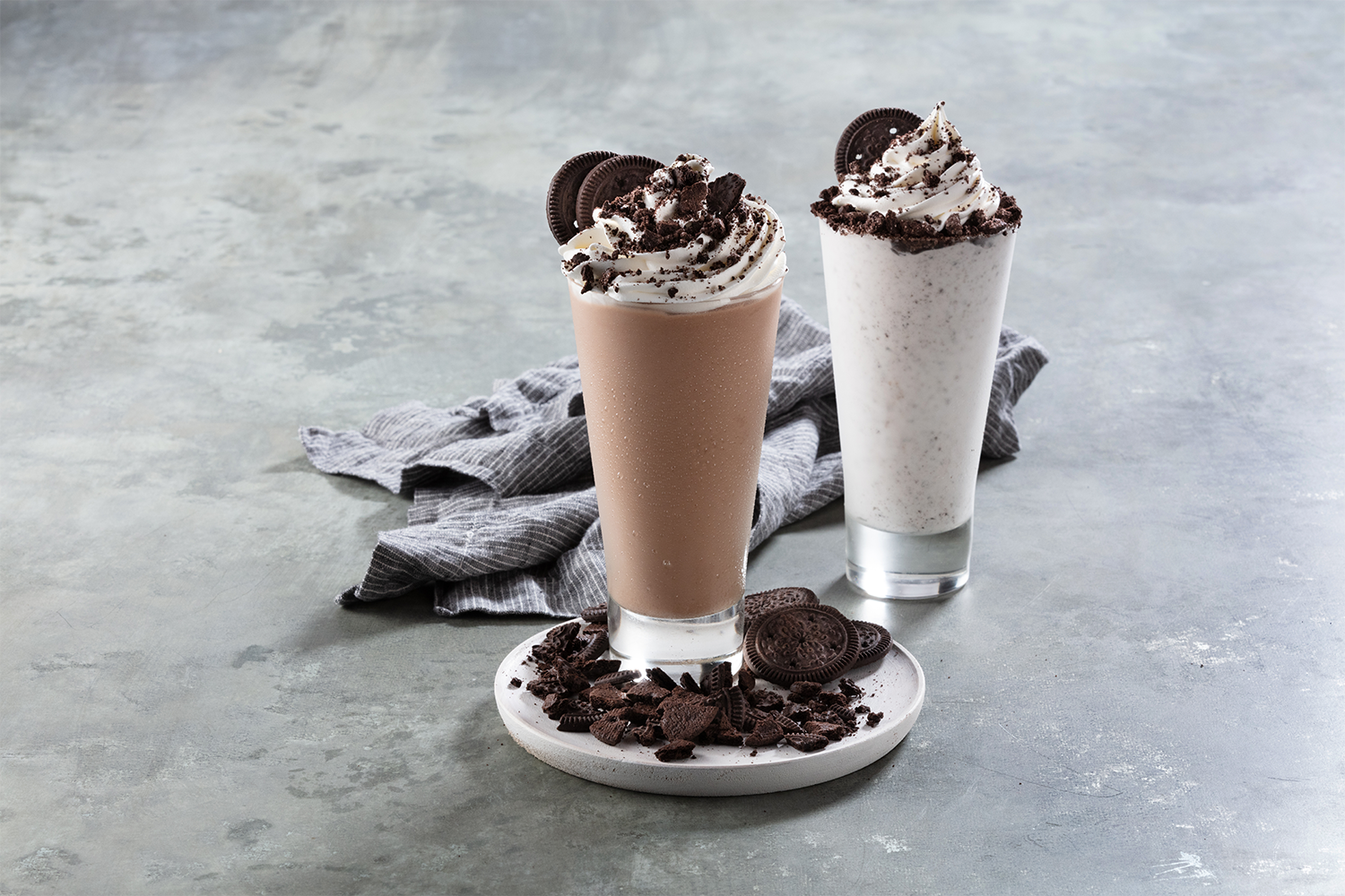 Two Oreo milkshakes topped with whipped cream and crushed cookies, one chocolate and one cookies and cream, placed on a white plate on a gray surface with a gray cloth nearby.