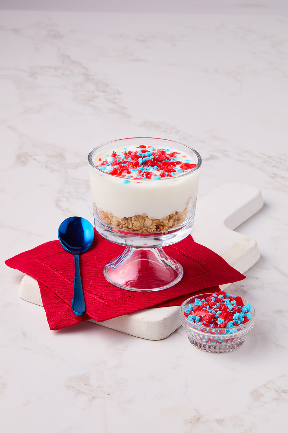 Layered dessert in a glass bowl with crushed graham crackers, white creamy yogurt, and red, blue, and white sprinkles on top, placed on a white marble surface with a red napkin, a blue spoon, and a small glass bowl of sprinkles nearby.