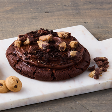 Close-up of a chocolate chip cookie with chunks of chocolate and nuts, on a white marble cutting board with mini cookies beside it.