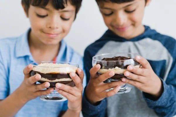 Two children holding dessert glasses filled with layered chocolate pudding, looking at them with smiles.