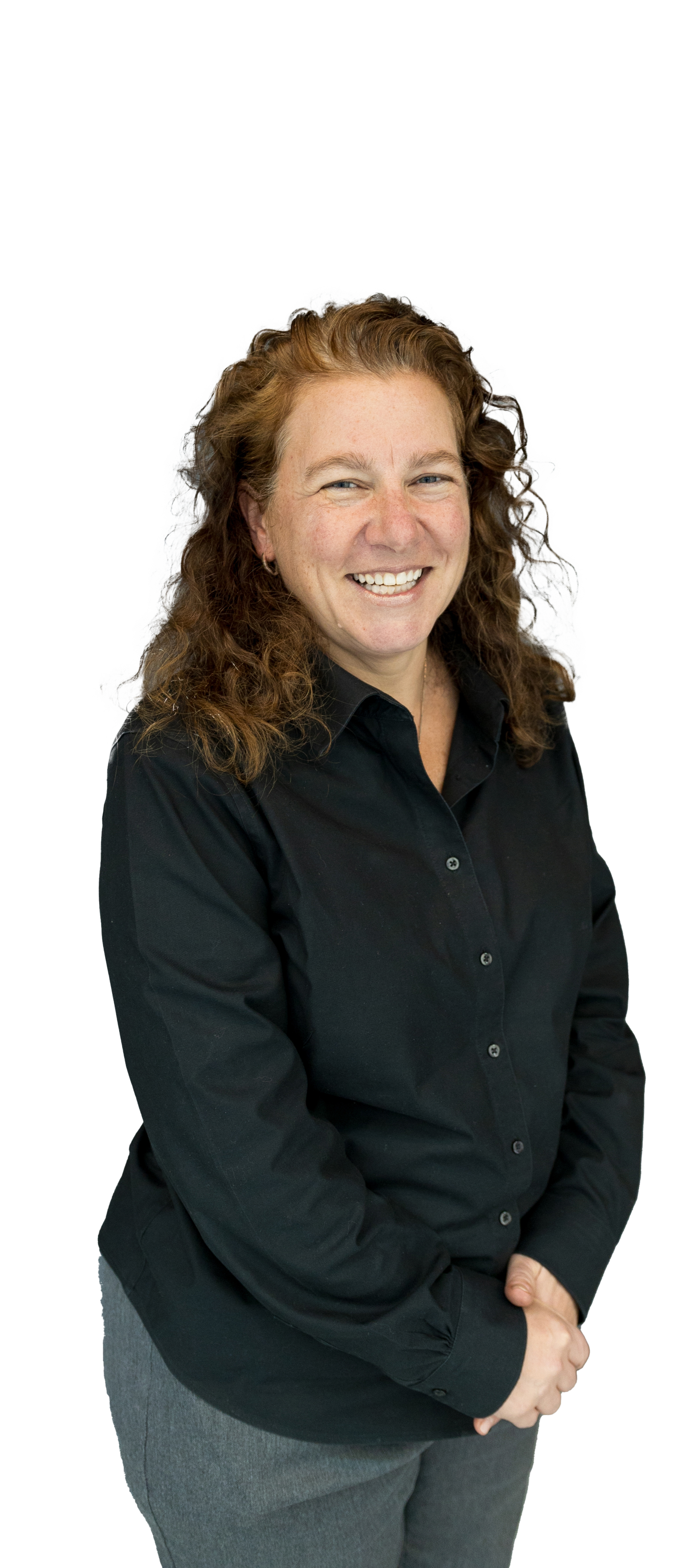 A woman with curly red hair smiling, wearing a black button-up shirt and gray pants, standing against a white background.