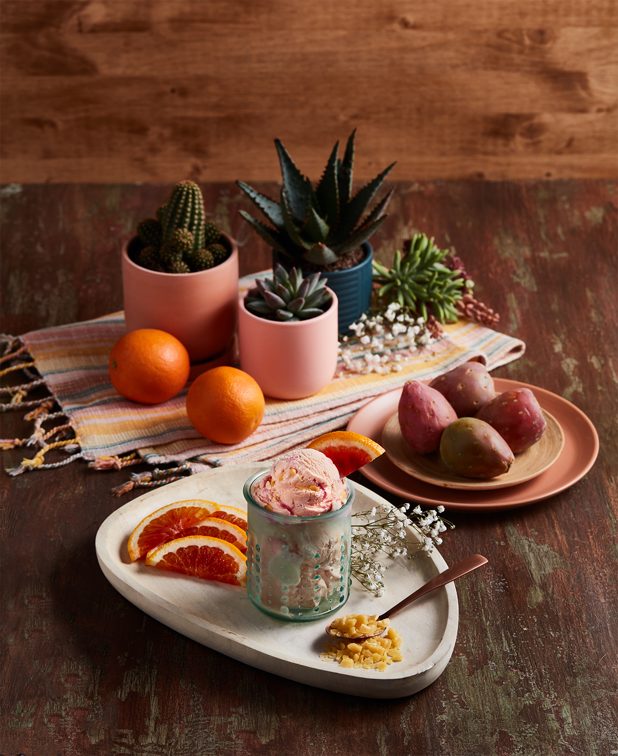 A dessert glass with pink-colored ice cream garnished with an orange wedge next to sliced blood oranges on a white serving board. Pink and peach plates with prickly pears, a tray of oranges, and potted succulents on a colorful striped cloth on a rustic wooden table.