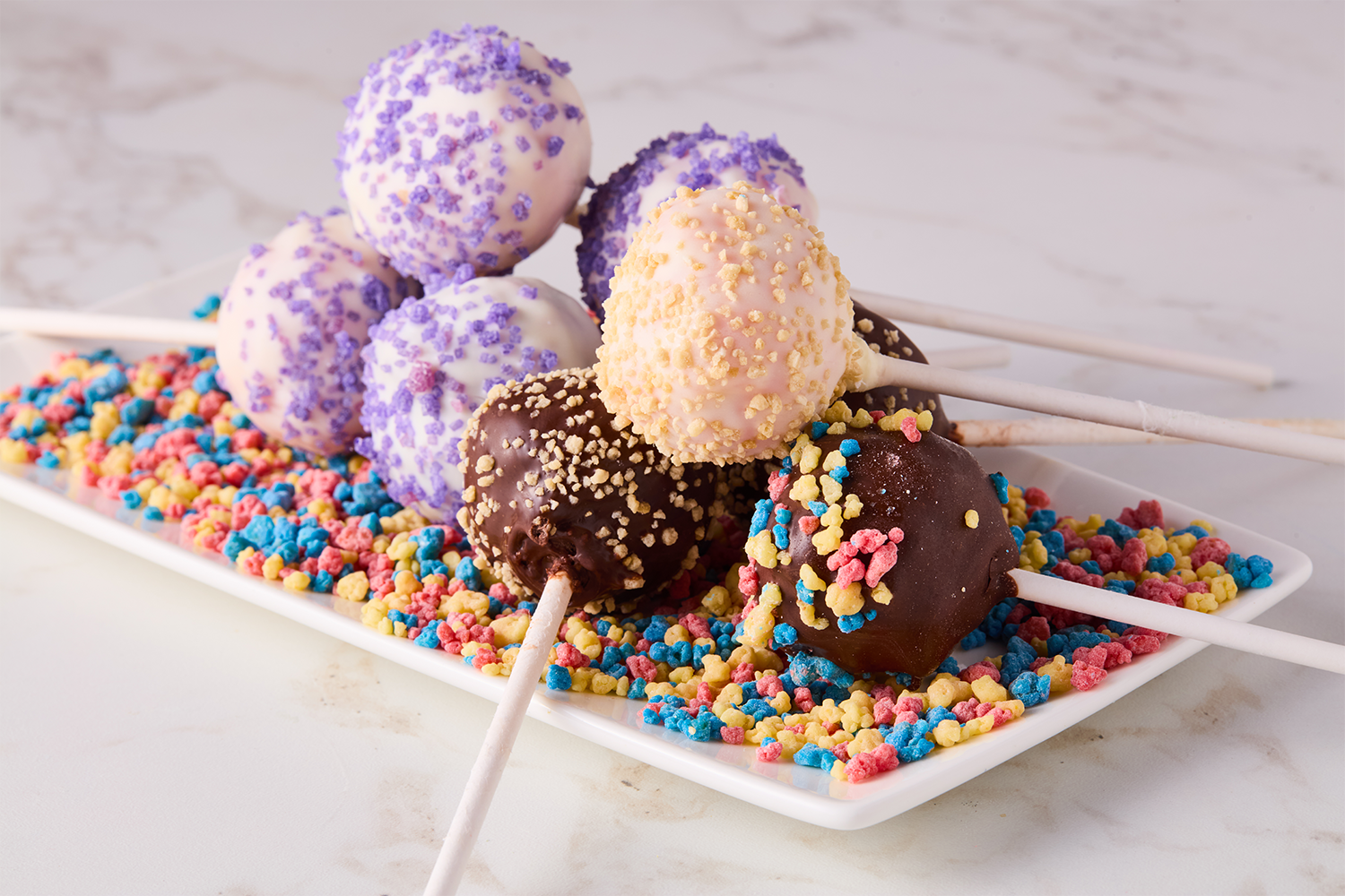 Assorted cake pops decorated with colorful sprinkles on a white rectangular plate.