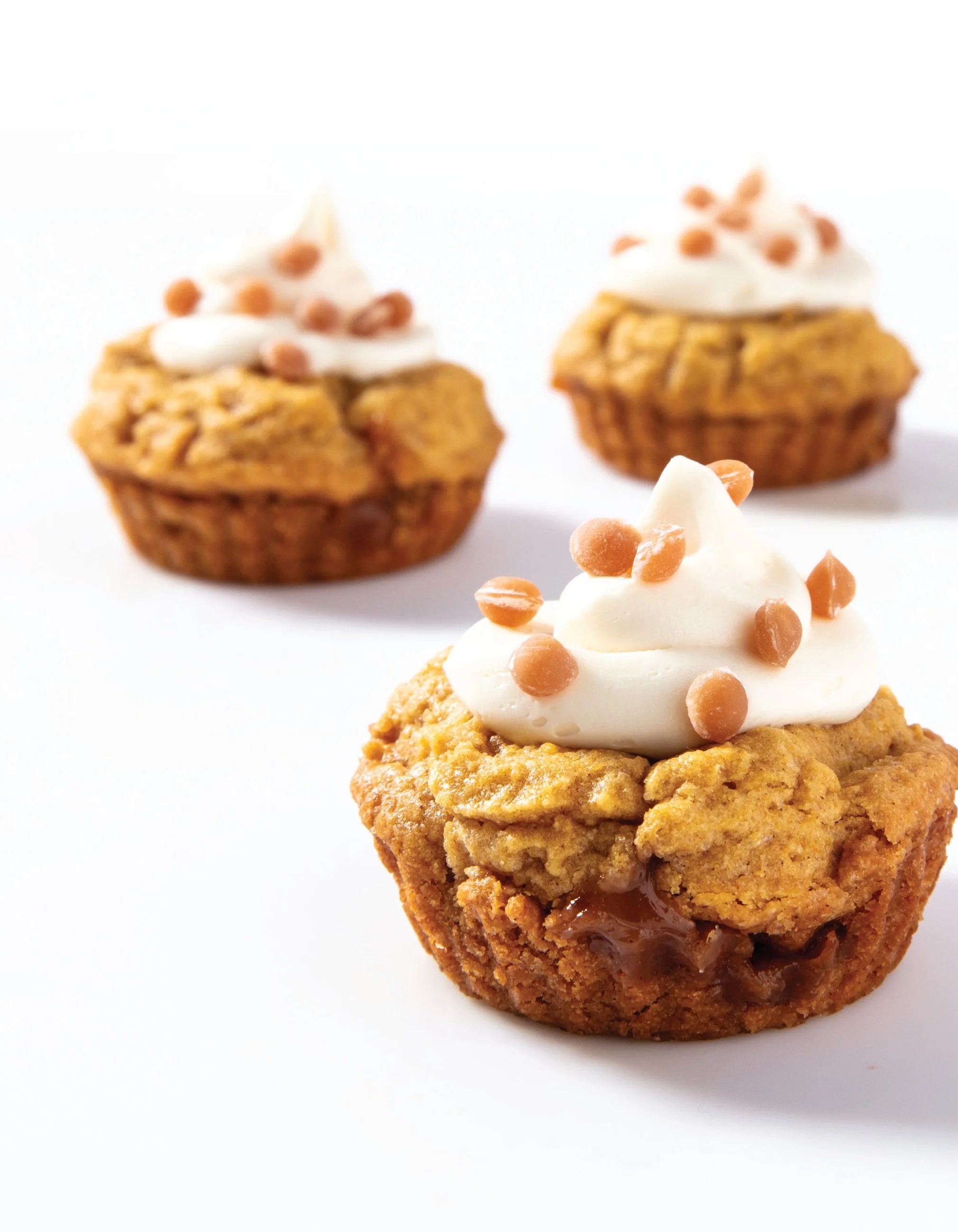 Three muffins with whipped cream and caramel chips on a white background, closest muffin showing a caramel filling.