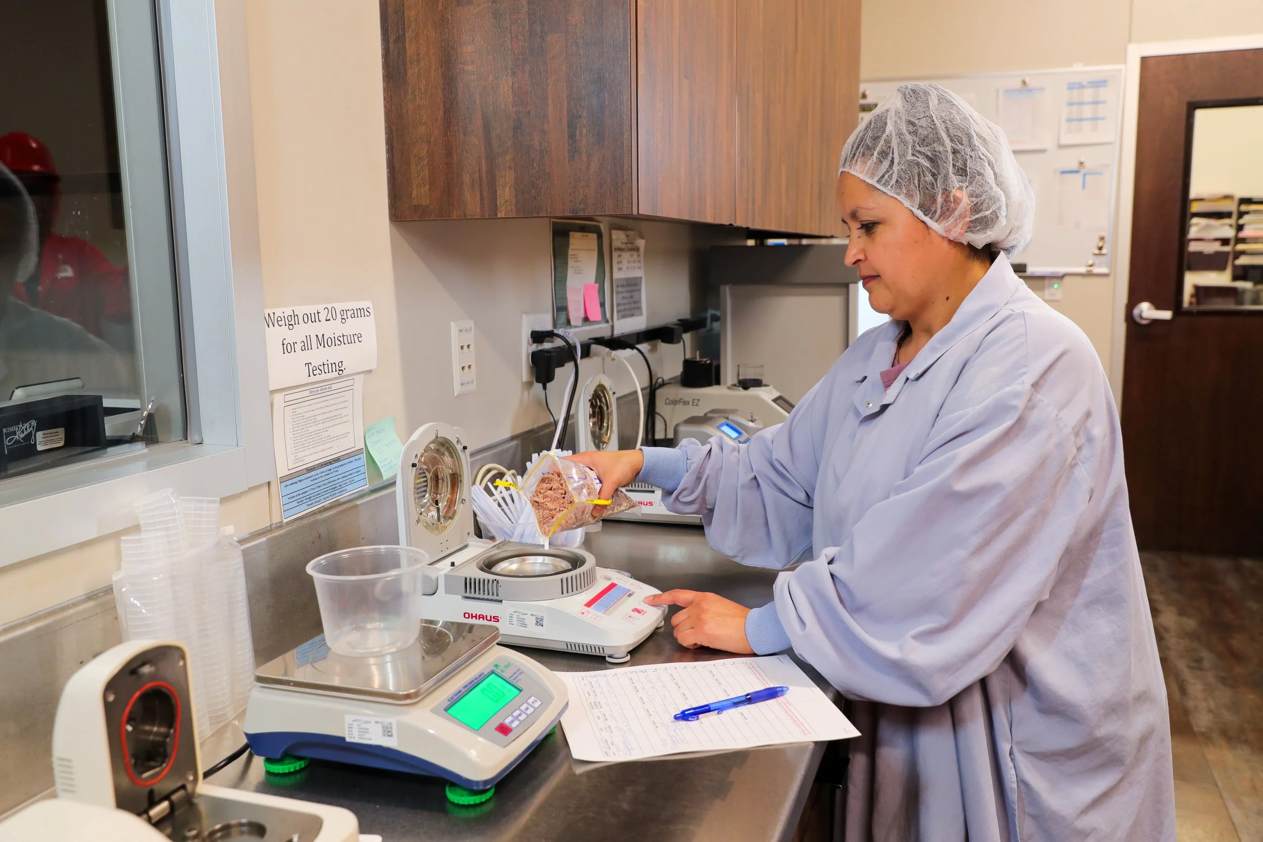 A woman wearing a lab coat and a hairnet weighs a piece of meat in a laboratory or clinical setting, with various laboratory equipment and paperwork on the counter.
