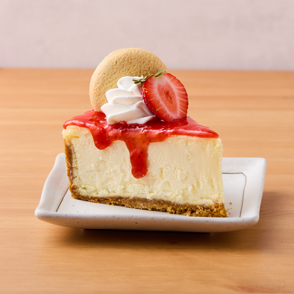 Slice of cheesecake with strawberry topping, whipped cream, a strawberry half, a round cookie, on a white plate.