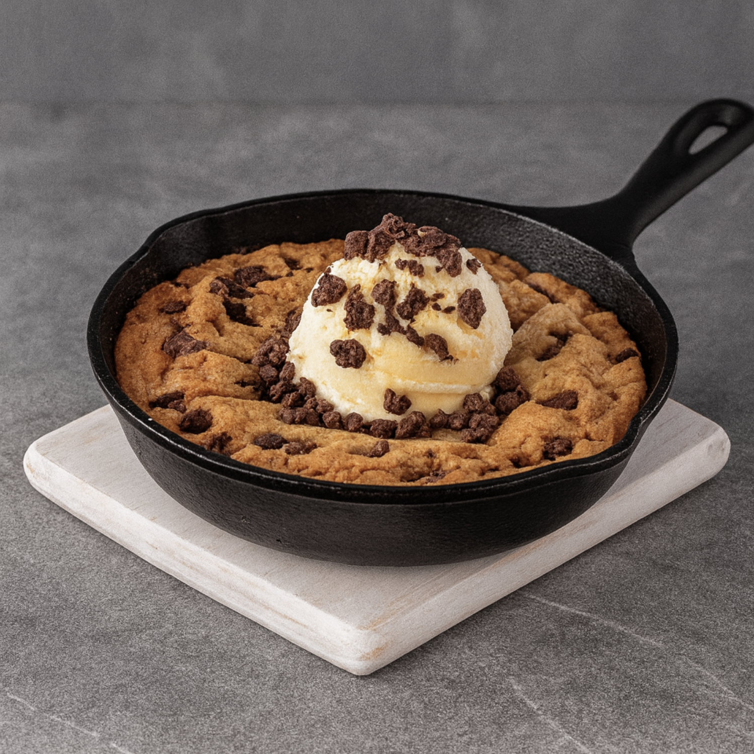 Cast iron skillet with chocolate chip cookie topped with a scoop of vanilla ice cream and chocolate shavings, on a white wooden board on a gray surface.