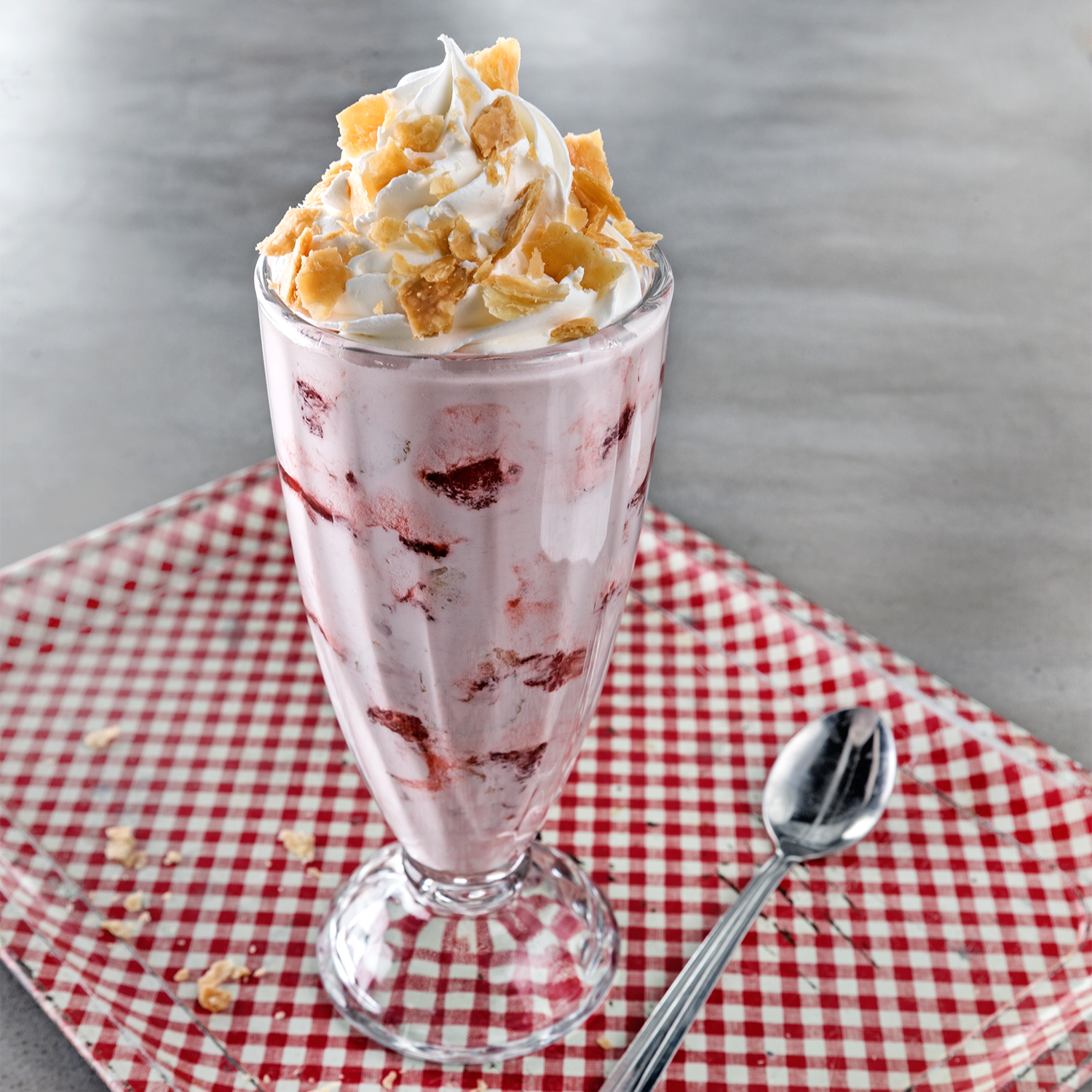 A tall glass of strawberry ice cream sundae topped with whipped cream and crushed cookies. The glass is placed on a red and white checked napkin with a spoon beside it.