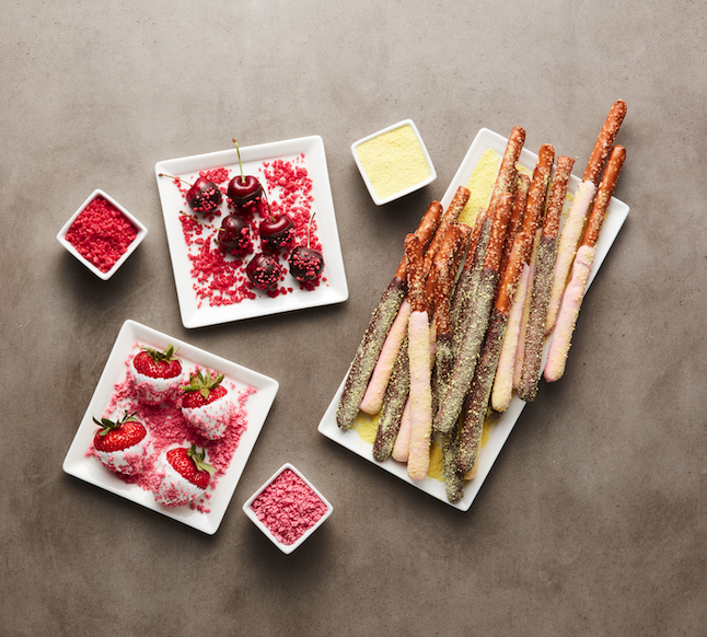 Assorted fruit and dessert items on white plates, including strawberries dipped in sugar, chocolate-covered strawberries, and churros with pink and yellow toppings, as well as small bowls of pink and red powdered toppings and a yellow dipping sauce.
