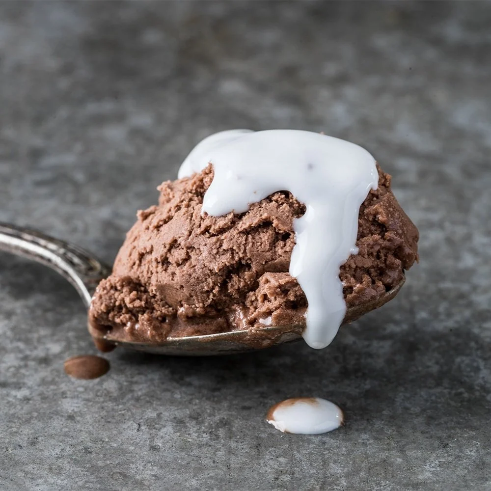 A scoop of chocolate ice cream with a spoon, topped with melting white cream or milk, on a gray surface.