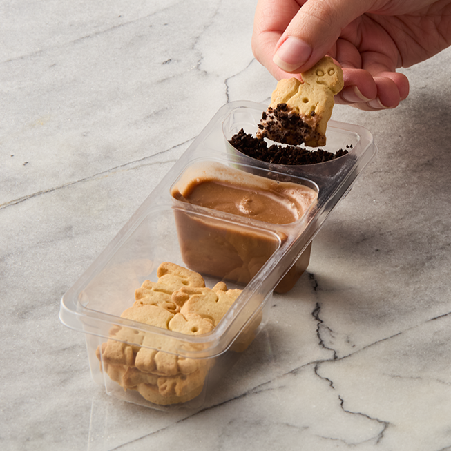 Person dipping a teddy bear-shaped cookie into a compartment of chocolate pudding in a three-section plastic container.