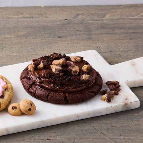 Chocolate cookie with chocolate chunks and cookie dough pieces on a marble cutting board, with small chocolate chip cookies beside it.