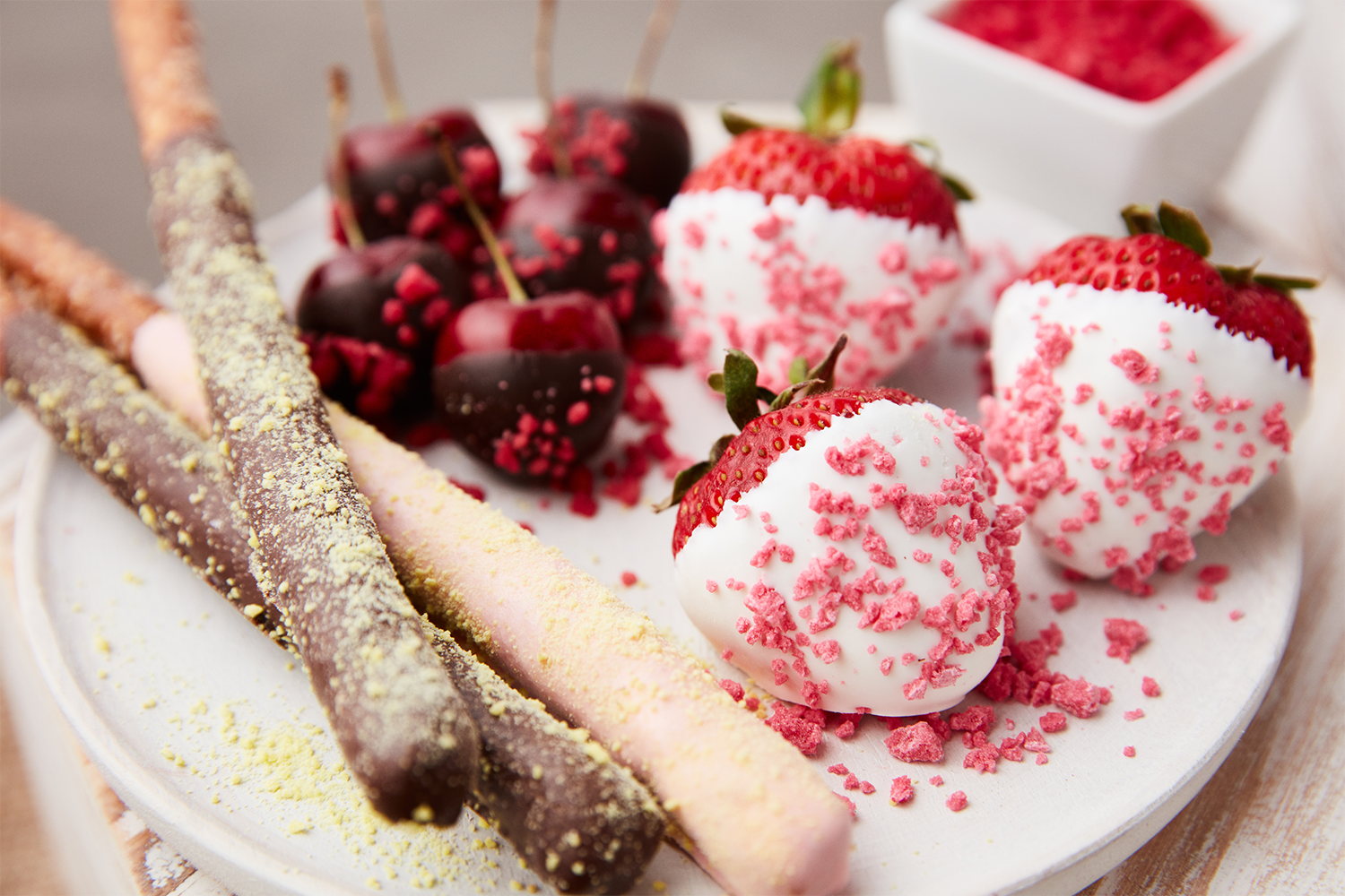 Assorted strawberries dipped in white and dark chocolate, pink and yellow-coated wafer sticks, and cherries on a white plate.