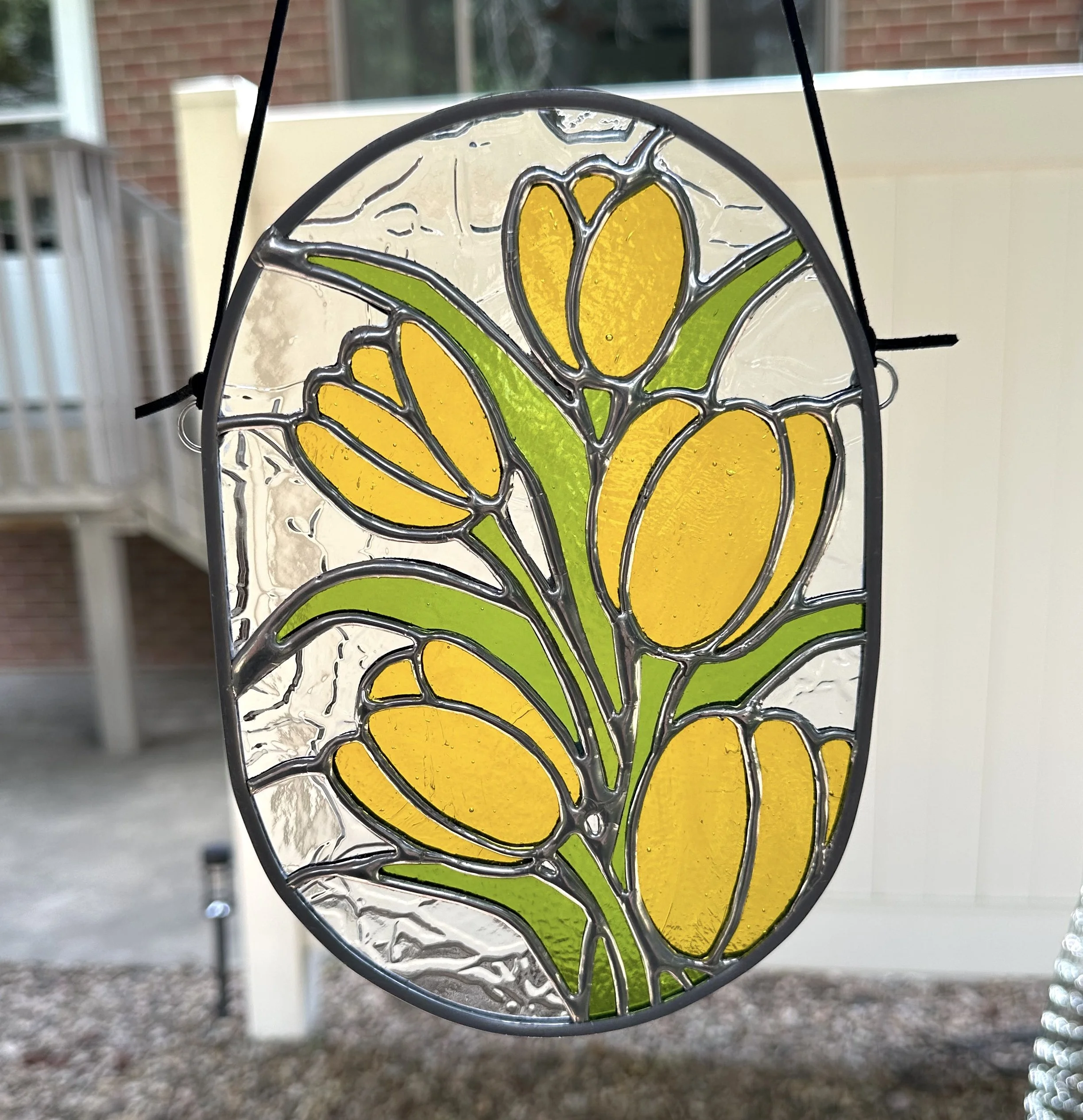 Colorful stained glass ornament with yellow tulips and green leaves, hanging outside.