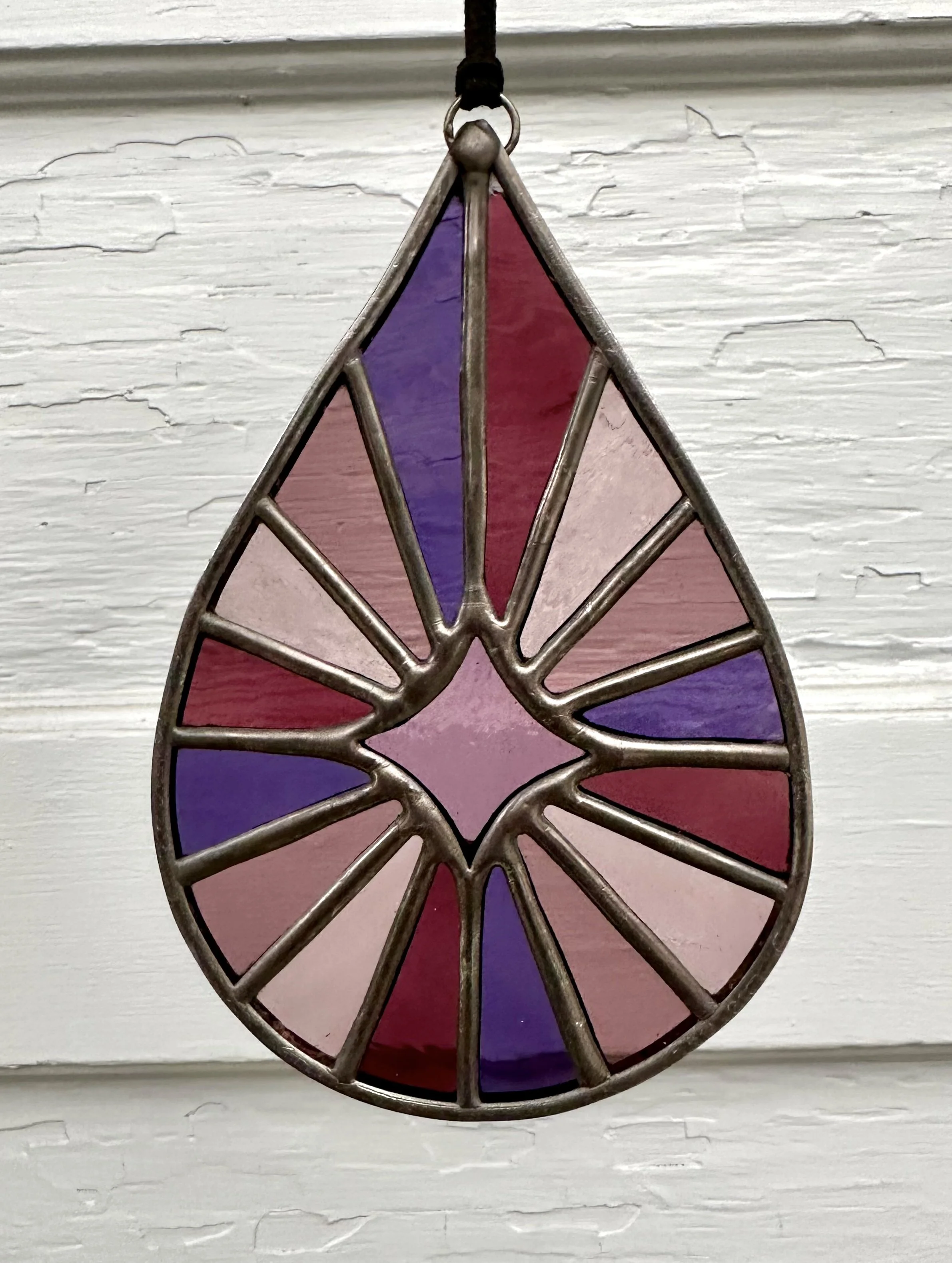 Colorful stained glass teardrop-shaped ornament hanging on a black string, with a design in shades of purple, pink, and red, against a textured white wooden background.