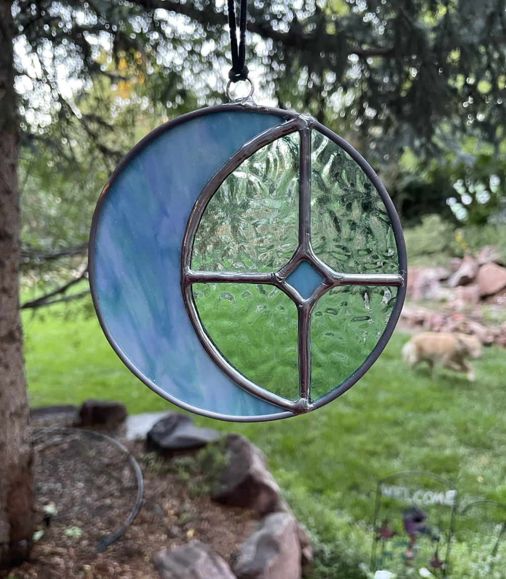 Colorful stained glass ornament with a moon and peace sign design hanging outdoors with trees and rocks in the background.
