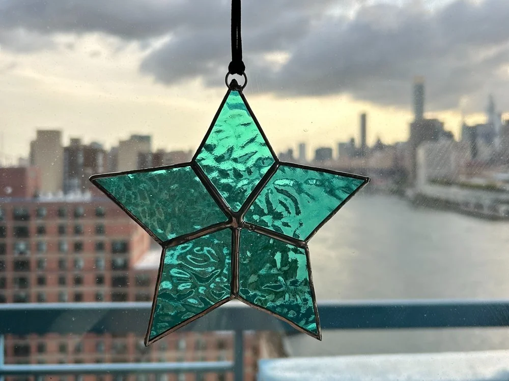 A stained glass star hanging in front of a window with a city skyline and river in the background.