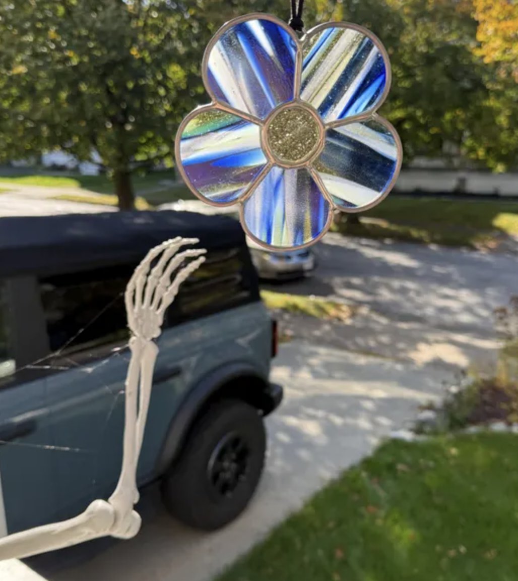 A decorative stained glass flower with blue and iridescent petals, held by a skeleton hand. In the background, there is a pickup truck on a suburban street with trees and a house.