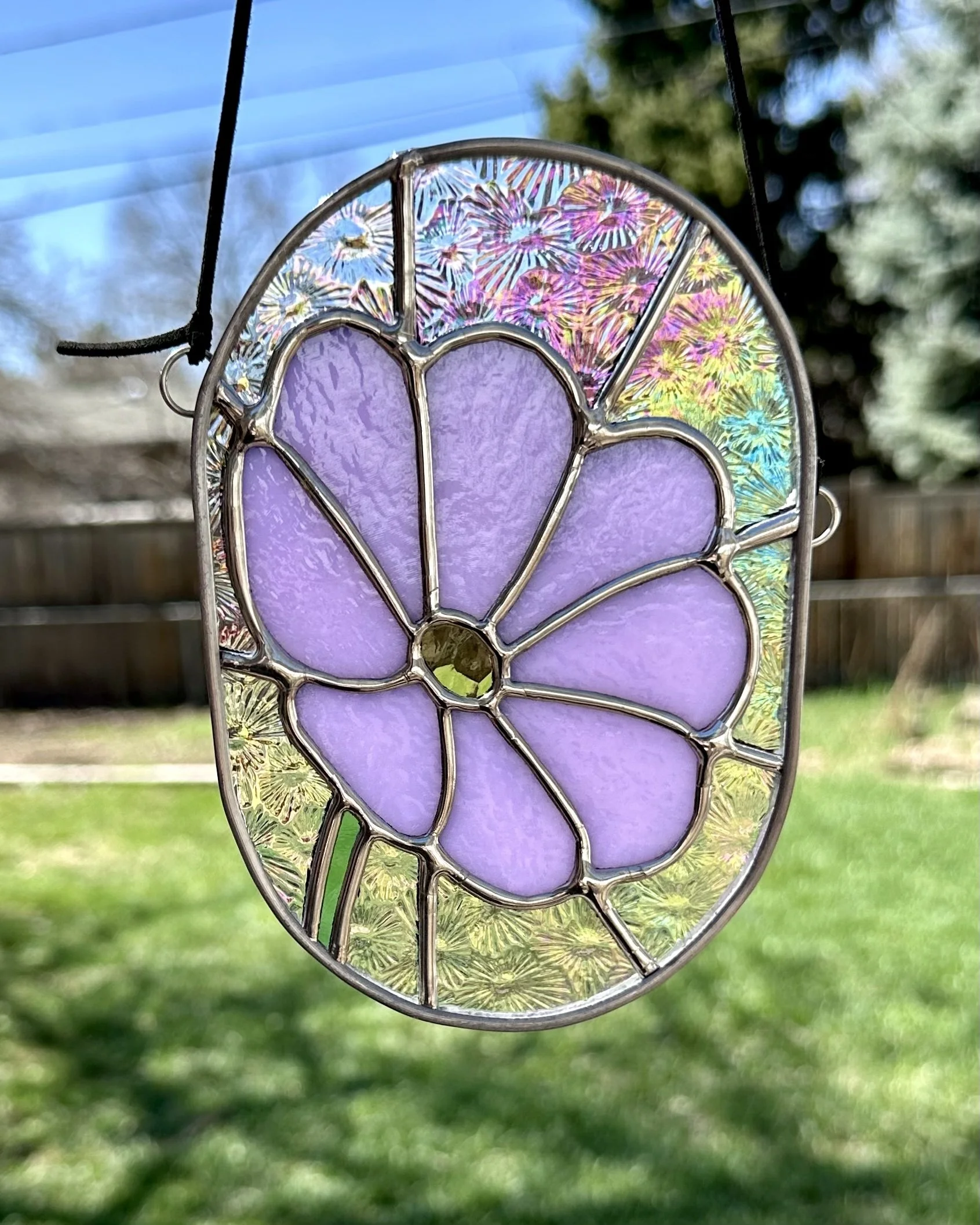 Colorful stained glass oval ornament with purple flower petals and a yellow and green background pattern, hanging outdoors with a blue sky and trees in the background.