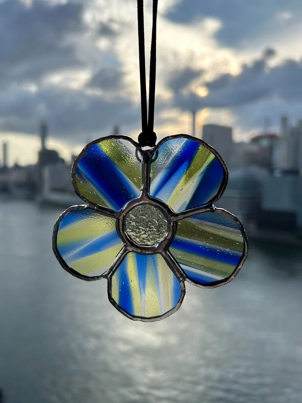 Stained glass flower ornament hanging outdoors during the daytime with a city skyline and cloudy sky in the background.