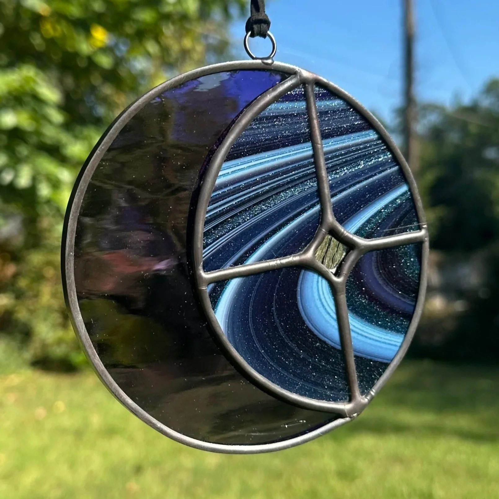 A stained glass suncatcher featuring a blue galaxy design and a peace sign in the center, hanging outdoors with trees and a blue sky in the background.