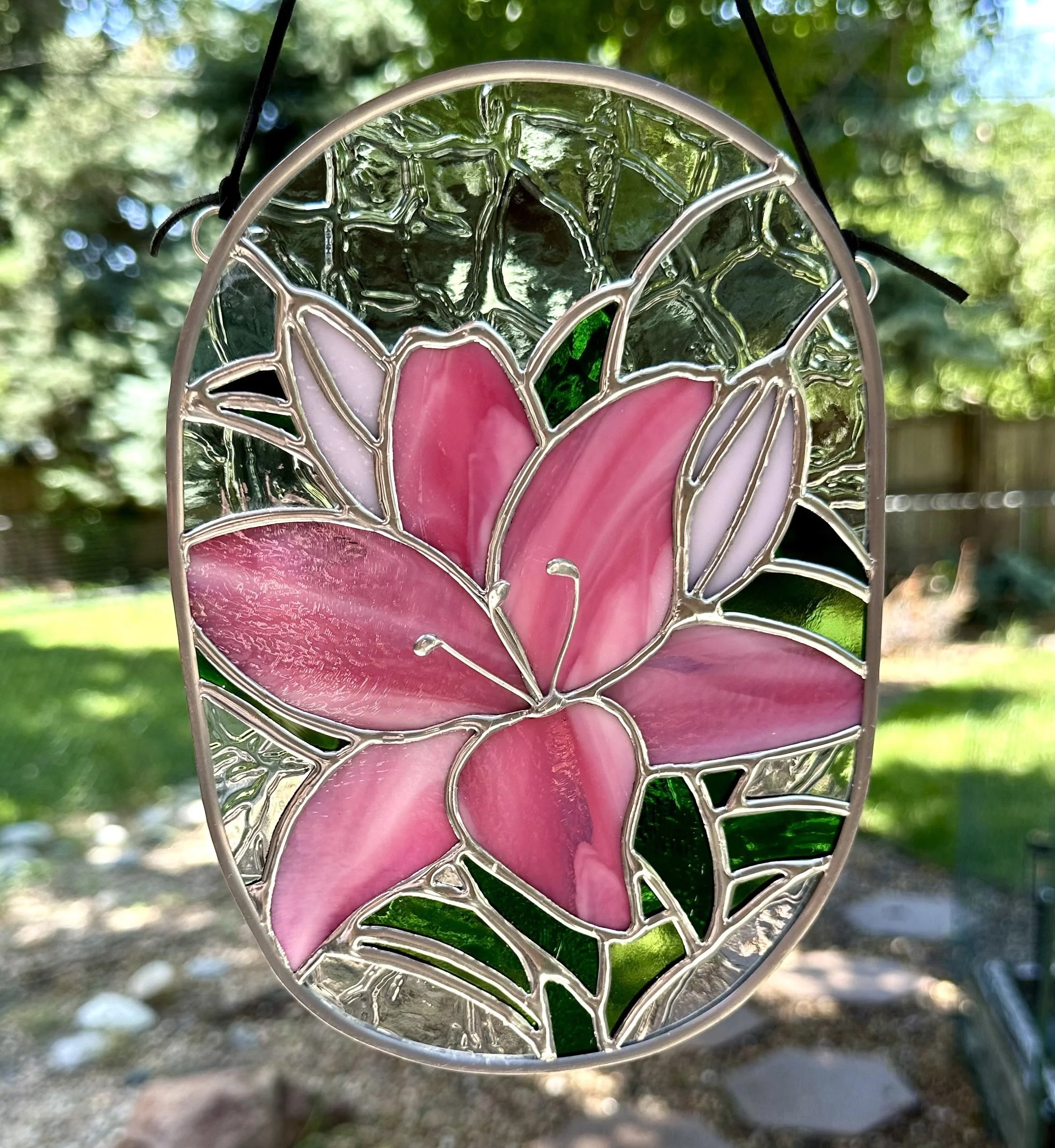 Stained glass art with pink lily flower and green leaves, hanging outdoors.