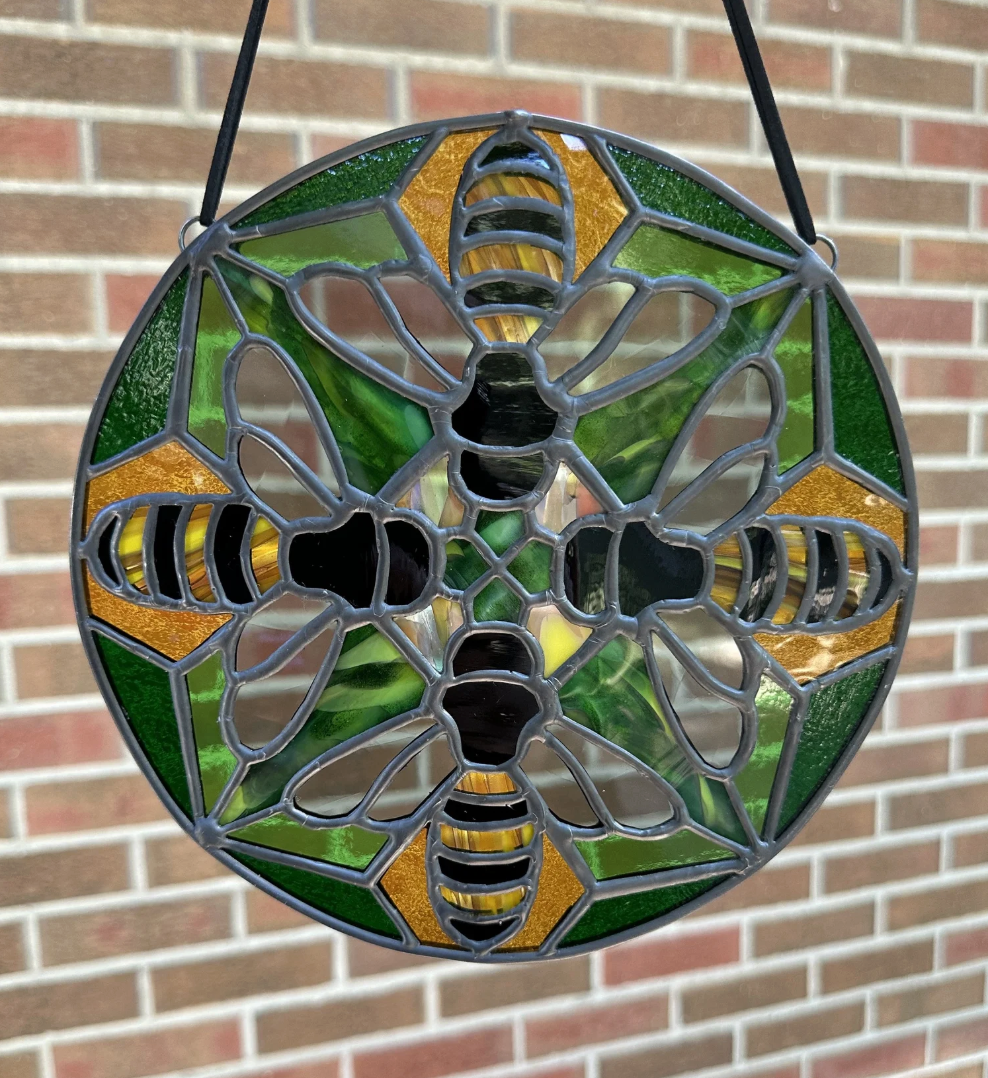 Stained glass ornament with a bee design, featuring yellow and black striped bees with green and yellow backgrounds, hanging in front of a brick wall.