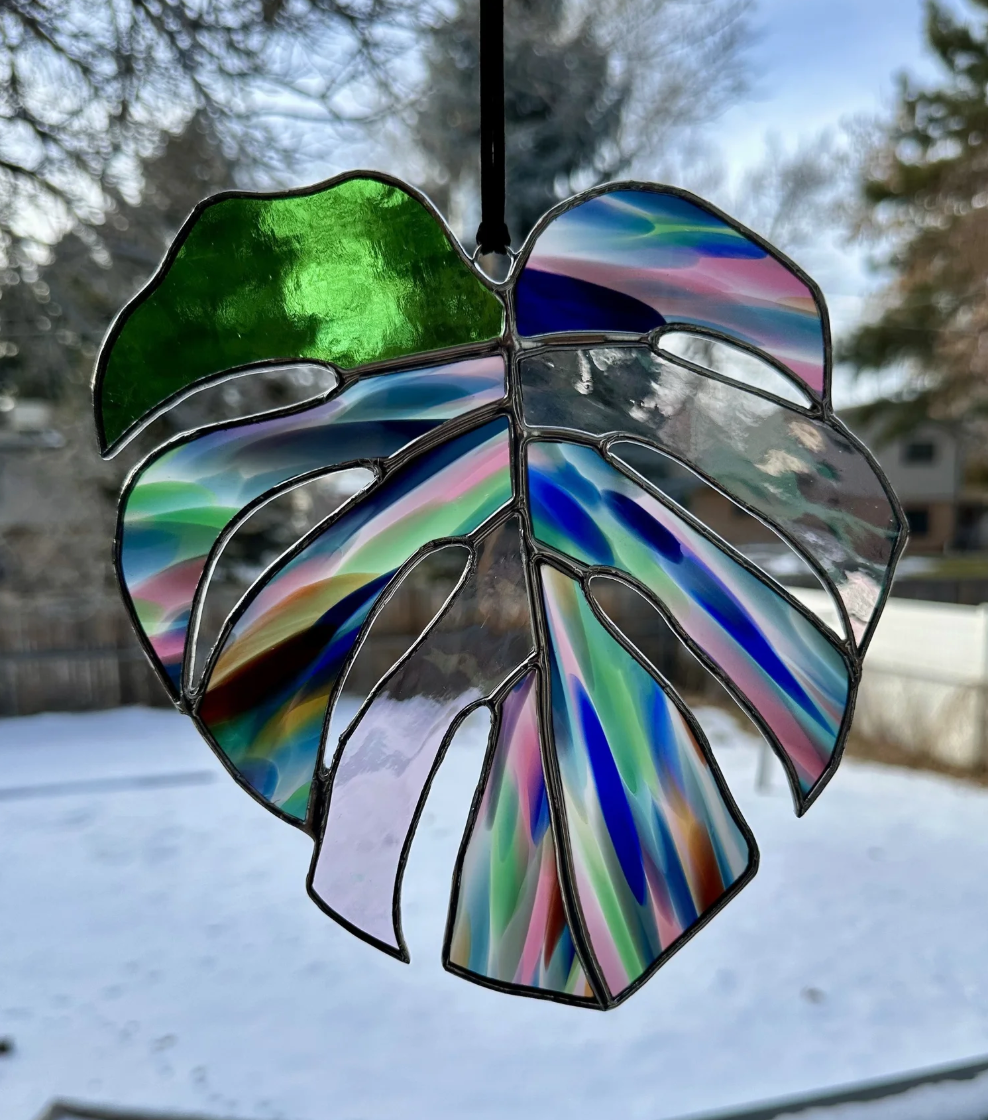 Colorful stained glass hanging shaped like a Monstera leaf, with a snowy outdoor scene and trees in the background.