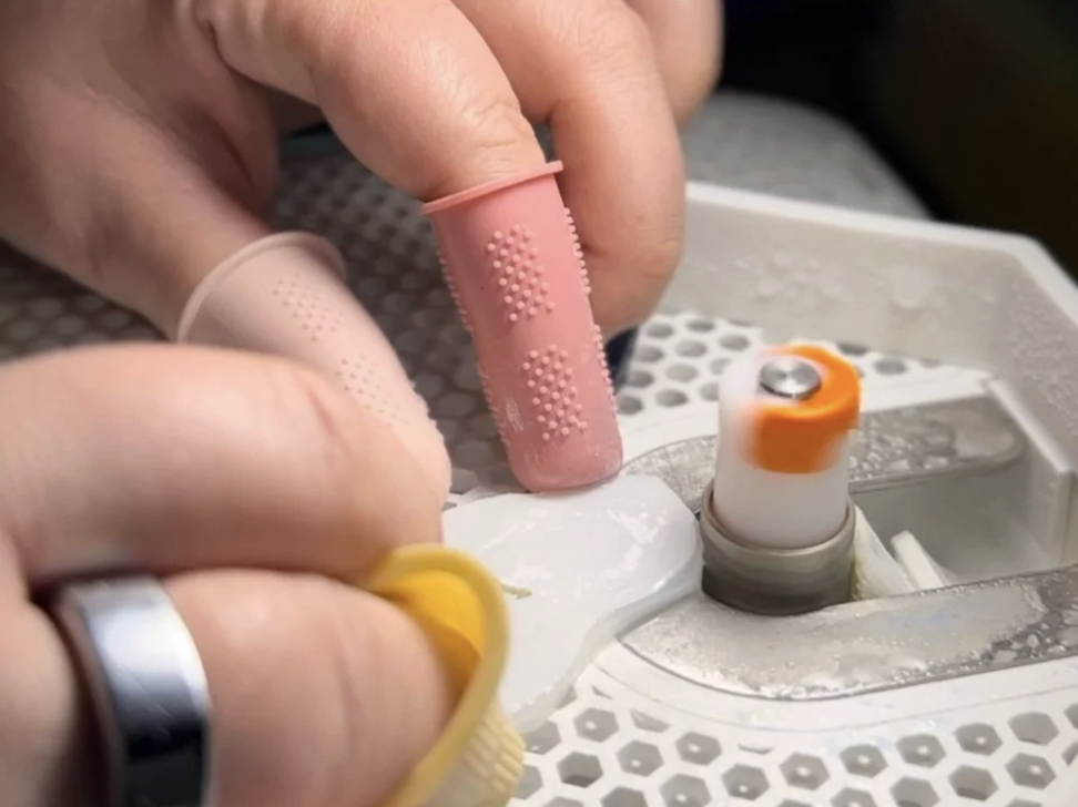 Close-up of a healthcare professional's hands using a pink finger stall and yellow tubing on a medical device, with medical equipment in the background.
