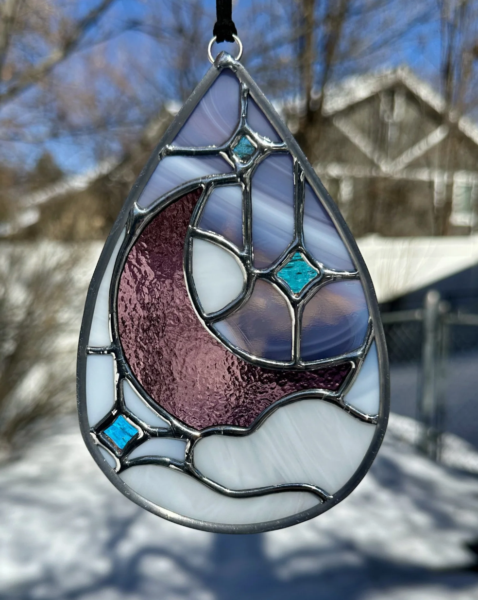 Stained glass pendant featuring a moon and star design with blue, purple, and white glass, hanging outdoors in a snowy background.