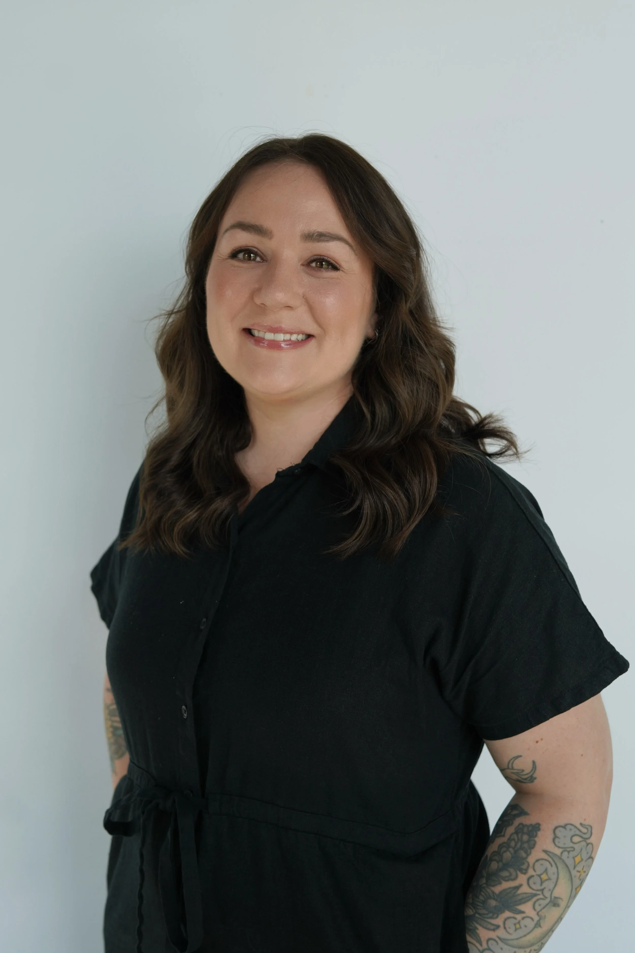 A woman with wavy brown hair and tattoos on her arms, wearing a black shirt, smiling at the camera against a plain light-colored background.