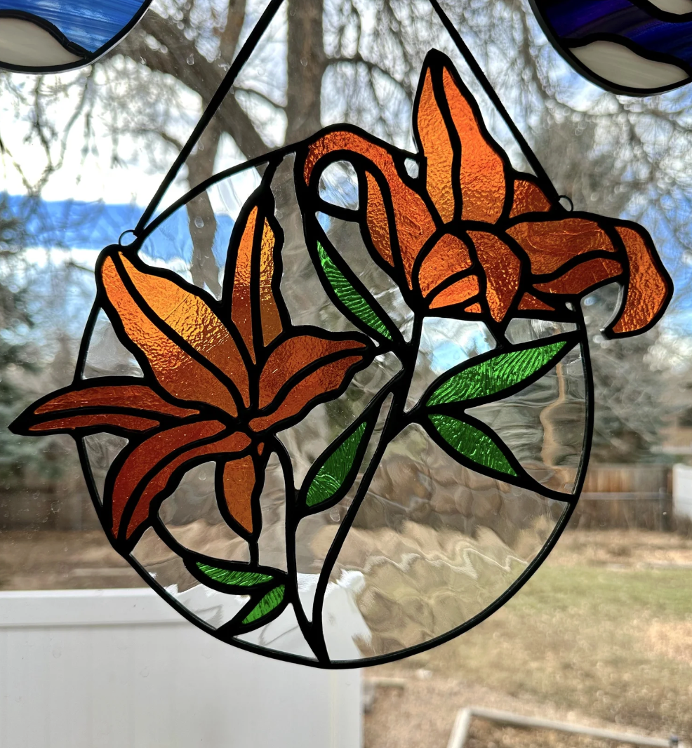 Stained glass hanging decoration featuring orange lilies and green leaves, with a blurred outdoor background.