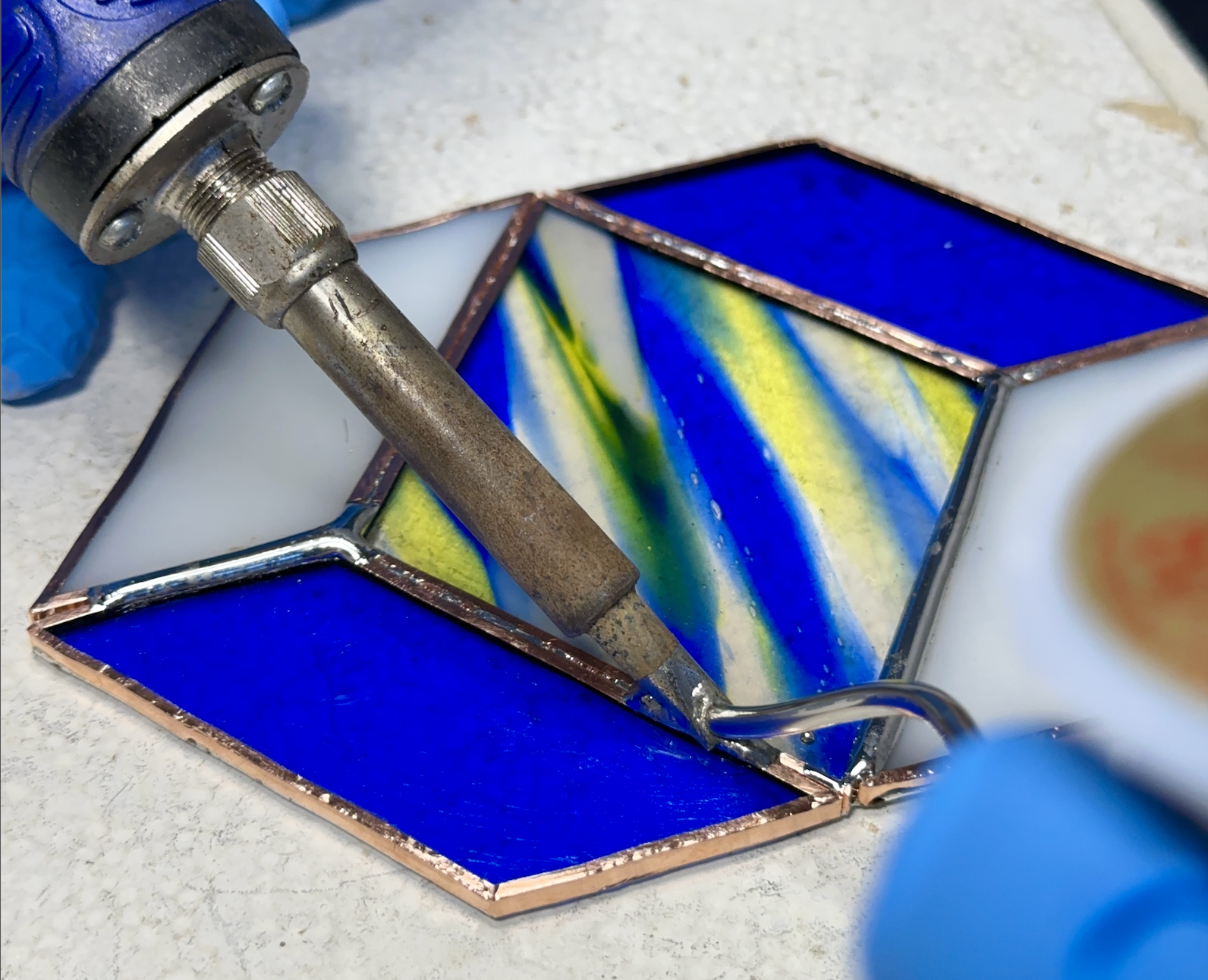 A stained glass piece with blue, green, yellow, and white colors being heated with a soldering iron.