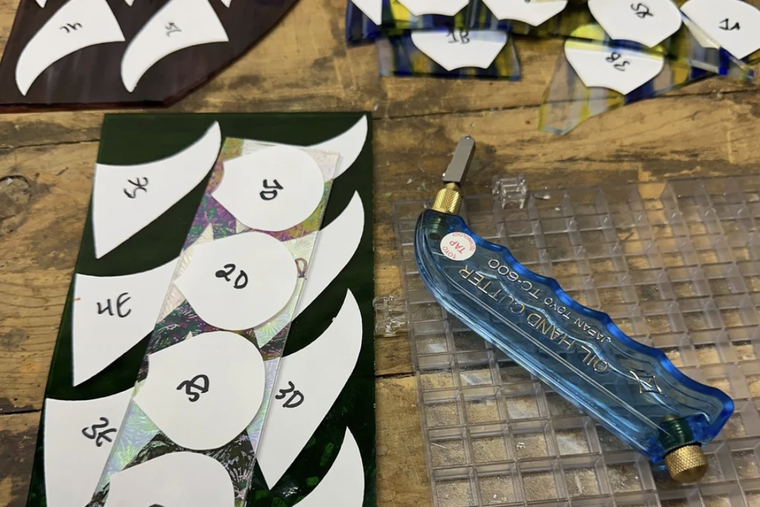 A craft project in progress with a green background sticker sheet featuring white oval cutouts labeled with different angles (30, 20, 60, 45, 35, 15, 10, 55, 5, 50, 40, 25). There is a precision utility knife with a blue handle and a clear plastic storage tray on a wooden surface.
