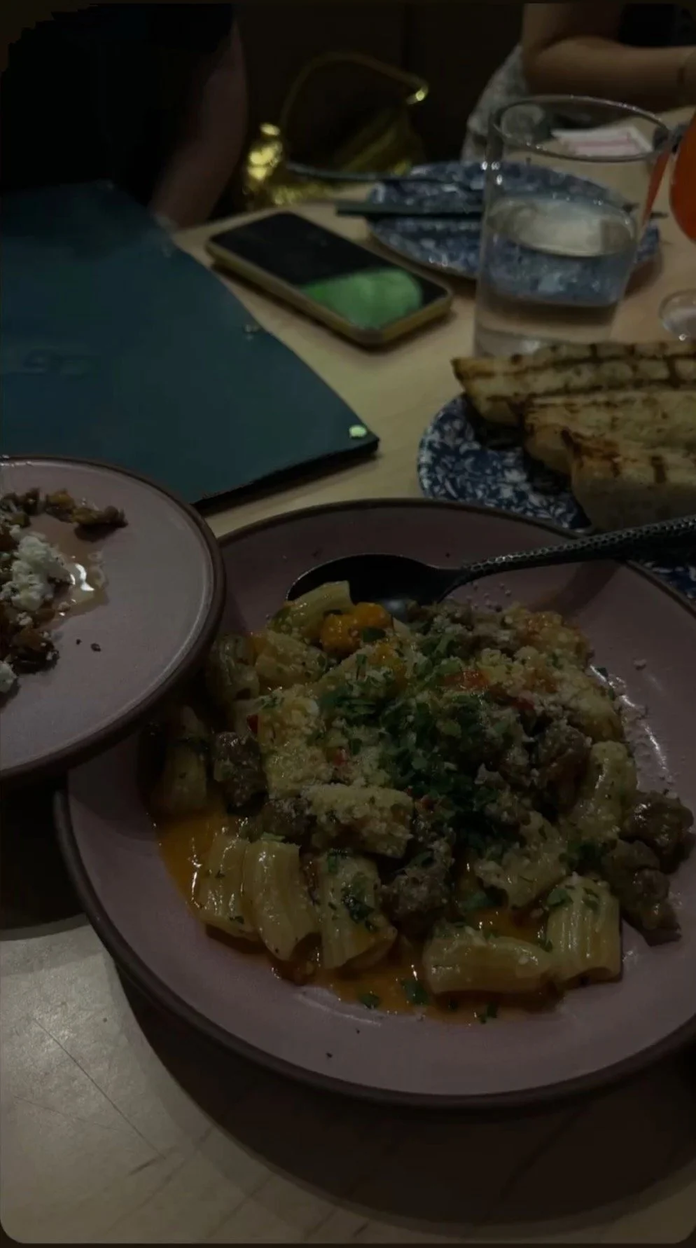 Plate of cooked pasta with meat and herbs, side of chopped grilled meat, water glasses, grilled tortillas, smartphone, notebook, and utensils on a dining table.