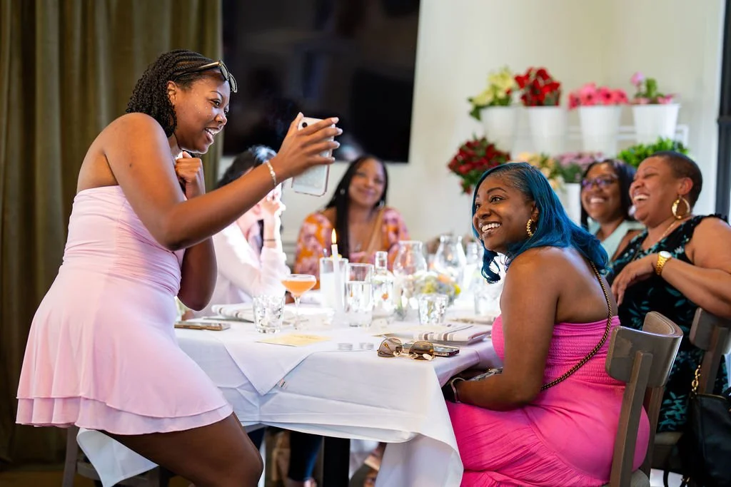 A woman taking a photo of a group of women sitting at a banquet table, all smiling and laughing, in a decorated indoor setting.