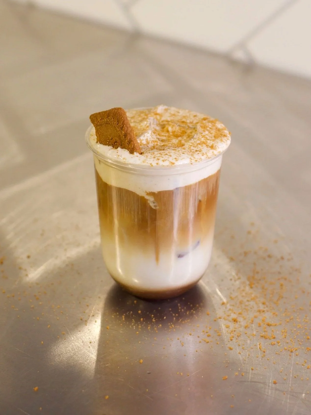 A glass of layered coffee drink topped with whipped cream, cinnamon, and a cookie, on a metallic surface with cinnamon powder sprinkled around.