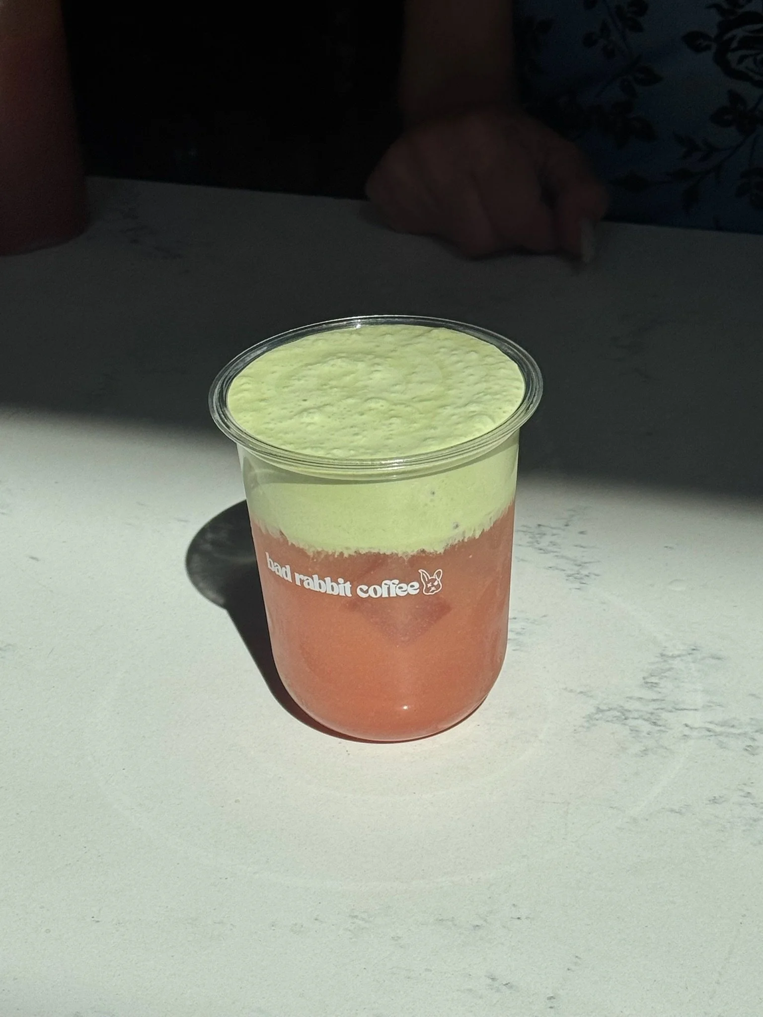 A cup of matcha latte with a gradient from green at the top to pink at the bottom, placed on a white surface with a faint gray marble pattern.