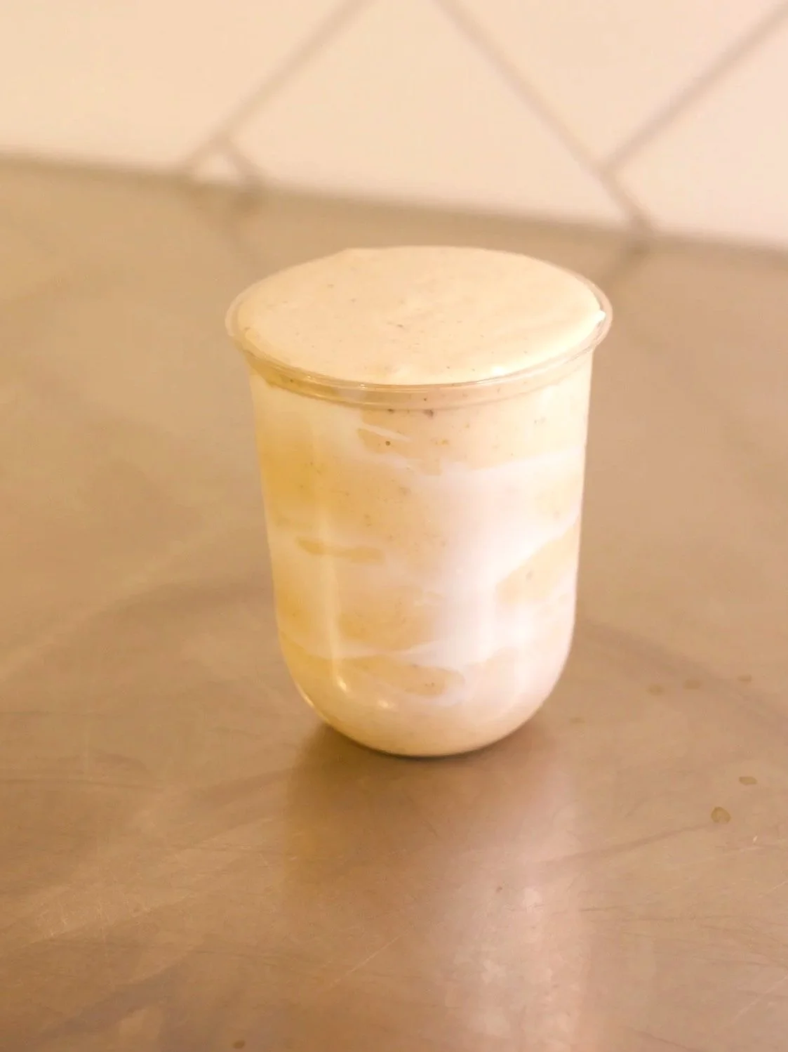 A small clear container filled with a creamy, beige-colored dessert, possibly ice cream or mousse, on a metallic surface with a plain background.