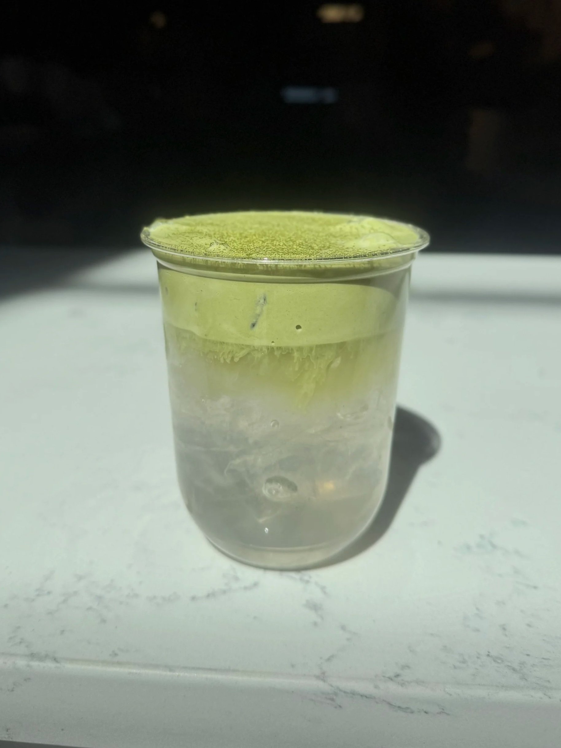 A clear plastic cup filled with a transparent liquid, with a greenish powder or substance evenly spread on the top surface.